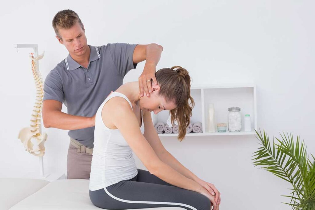 Adult practicing guided spine mobility exercises on a mat beside a treatment table.